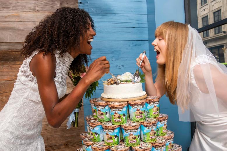 Two women laughing with each other while holdings spoons above an ice cream cake that is sitting on top of a stack of Ben & Jerry's ice cream tubs